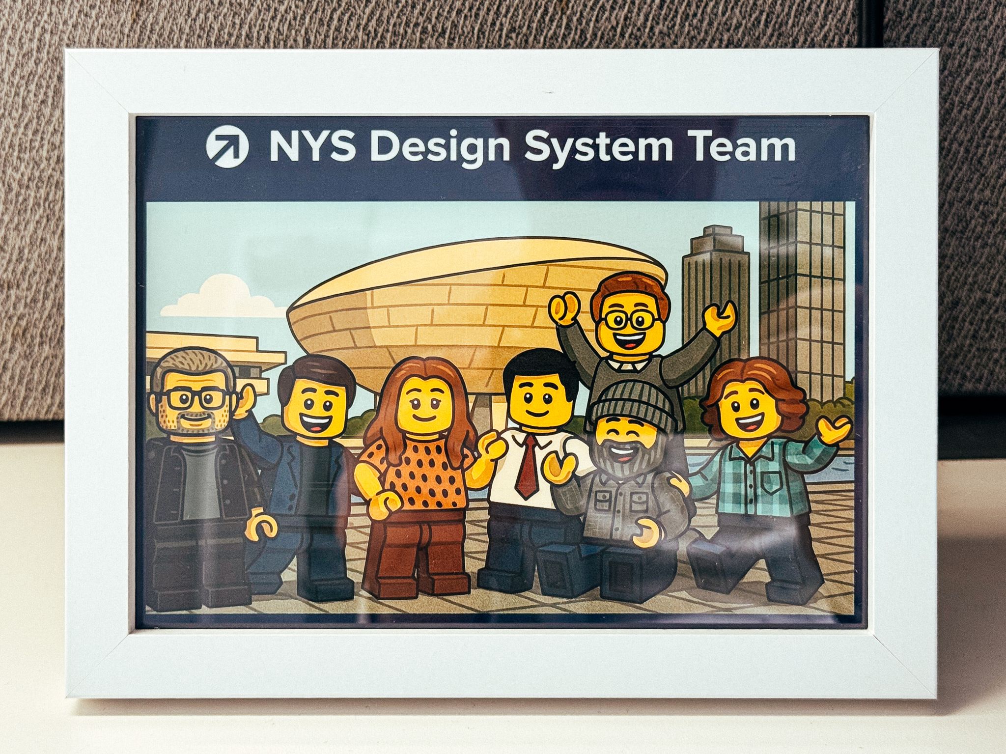 A framed illustration of the NYS Design System Team as Lego minifigures standing in front of The Egg at Empire State Plaza in Albany, NY.